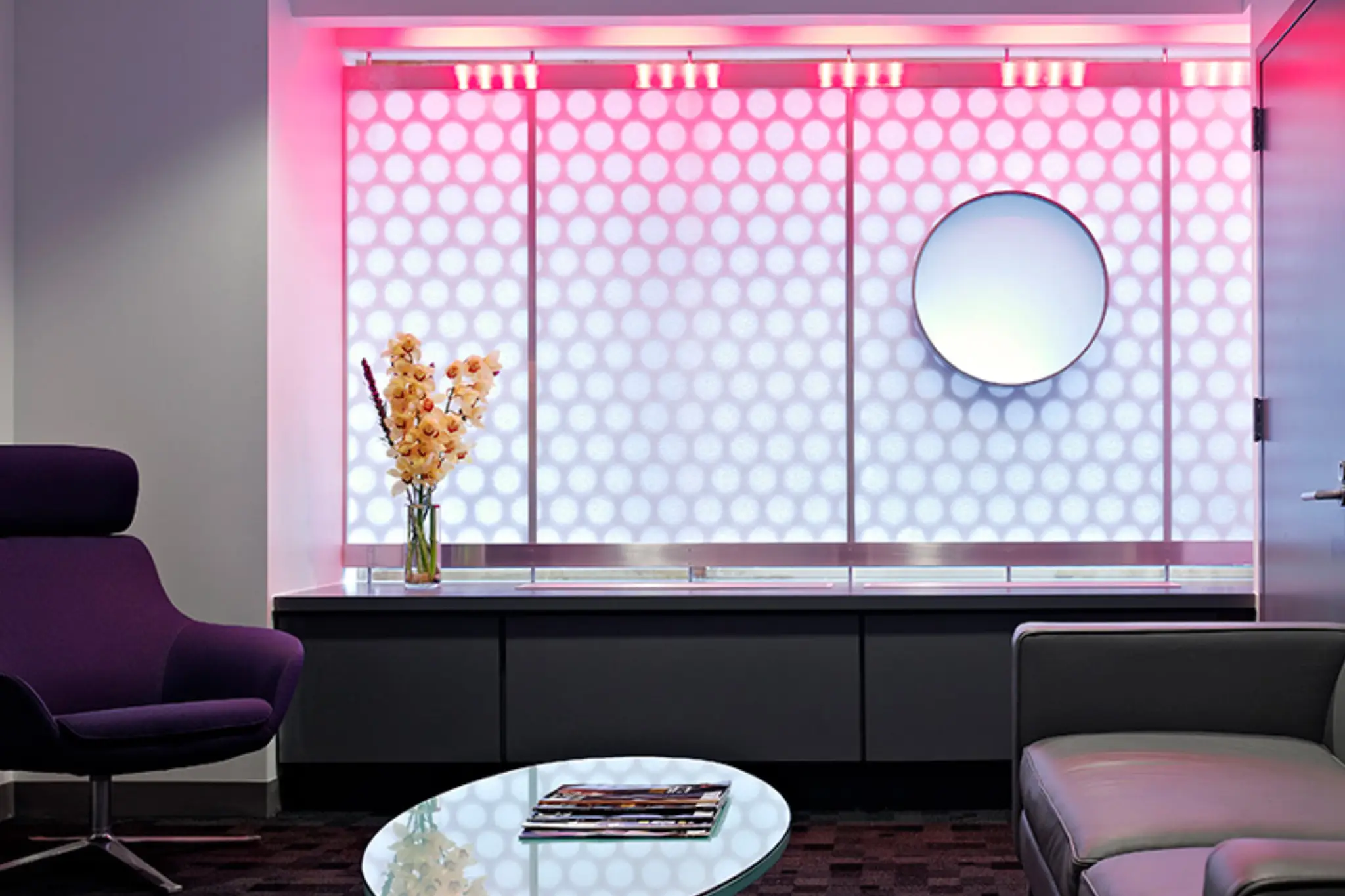 Modern lounge with purple chair, sleek sofa, circular mirror, and illuminated wall displaying patterned lighting. A vase of flowers on a glass table